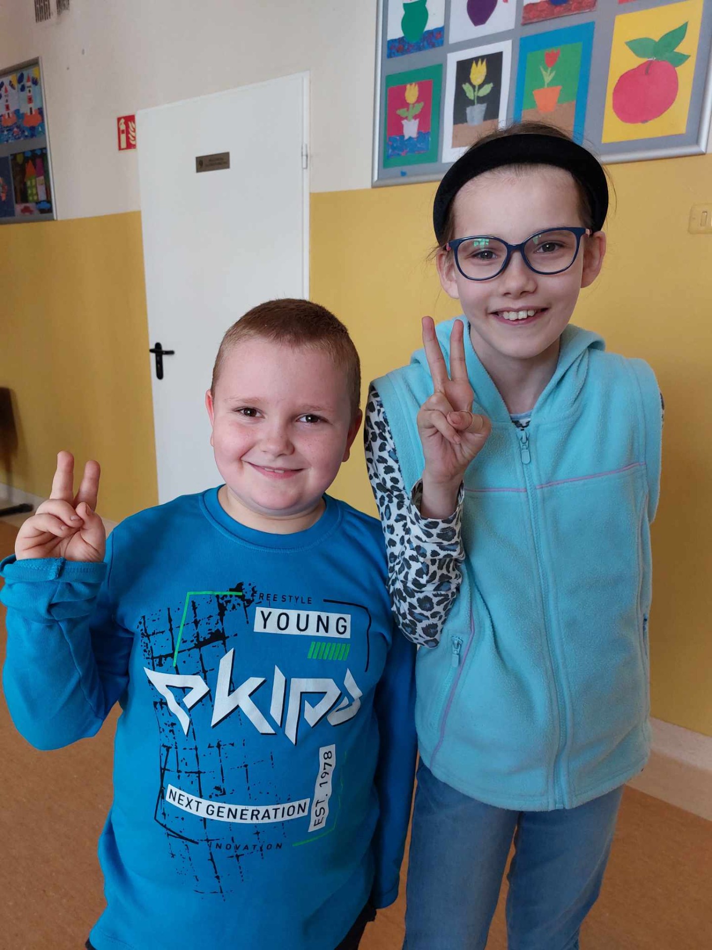 Chłopiec i dziewczynka stoją ubrani na niebiesko i pokazują literę V na palcach