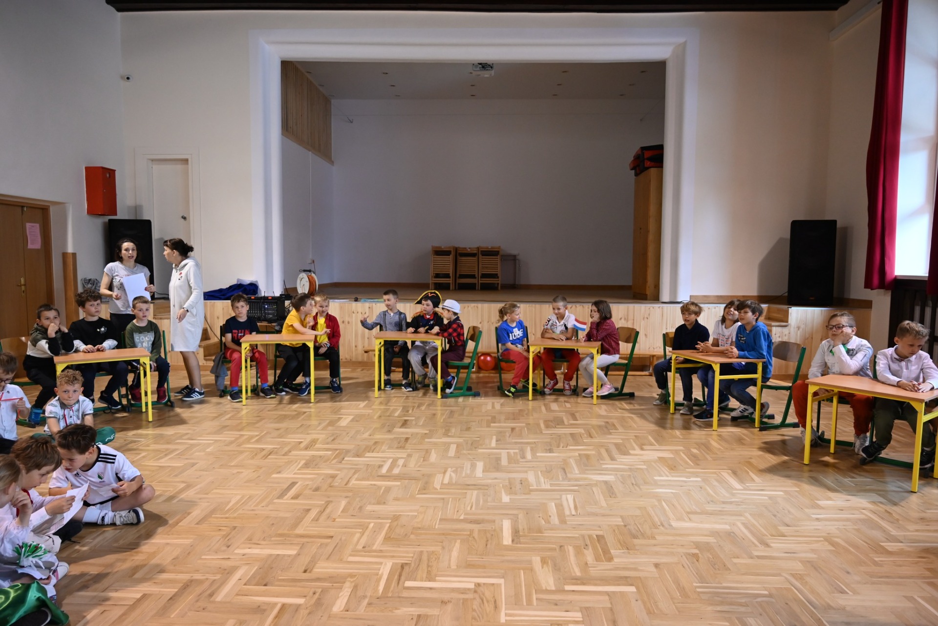 Liczna grupa dzieci siedząca przy stolikach podczas quizu wiedzy w sali gimnastycznej