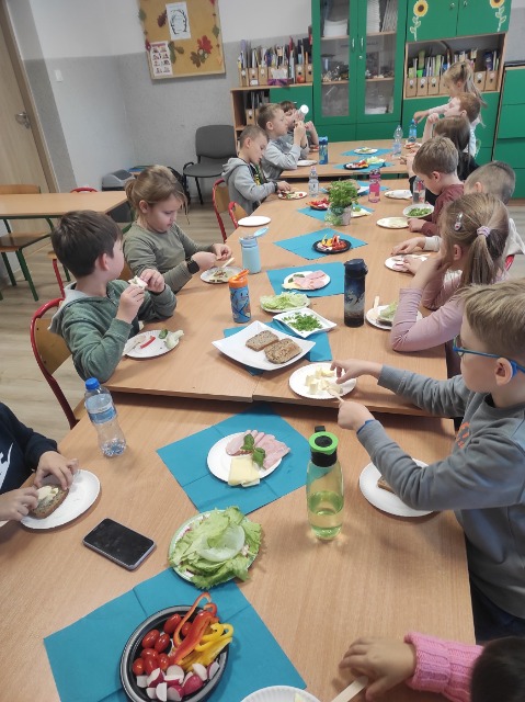 Dwunastu uczniów siedzi po dwóch stronach długiego stołu, który jest nakryty do przygotowania śniadania. Znajduje się na nim chleb, warzywa, masło, ser i wędlina