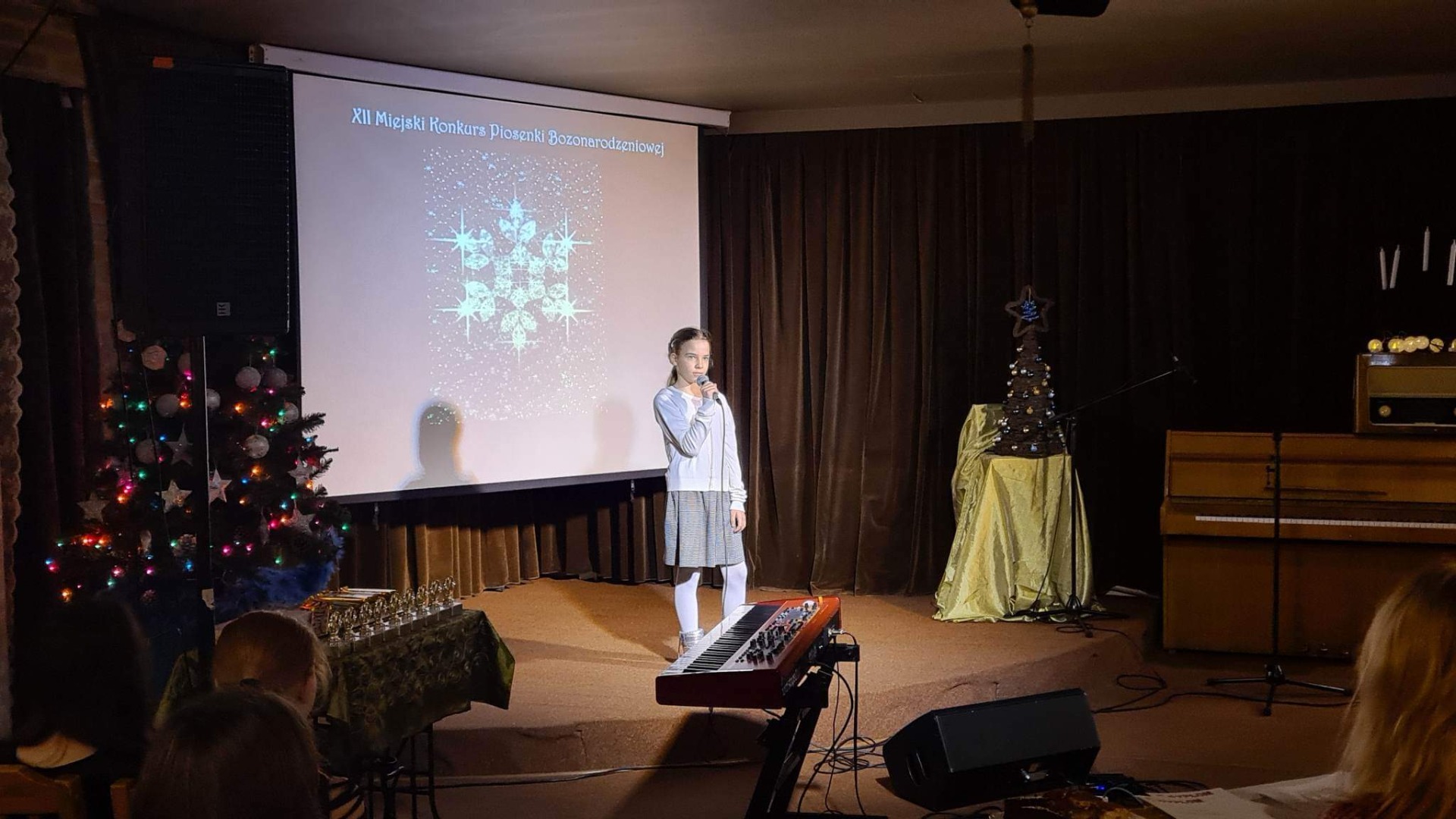 Dziewczynka w jasnym sweterku i spódnicy stoi na scenie. W ręku trzyma mikrofon. W tle widoczna dekoracja: prezentacja z motywami świątecznymi, choinka, pianino.