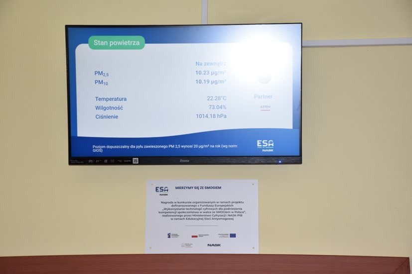 Monitor z wyświetlanymi danymi o jakości powietrza w otoczeniu szkoły i zamieszczona pod nim tabliczka z napisem ESA - Mierzymy się ze smogiem.