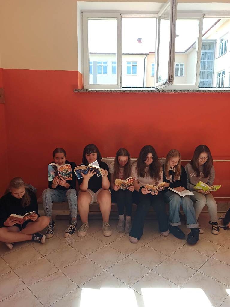 Zdjęcie przedstawia dziewczynki z klasy piątej. Sześć  siedzi na niskiej ławce w szatni, a jedna na posadzce. Wszystkie czytają książki. W tle za uczennicami znajduje się pomalowana na pomarańczowo ściana, a wyżej otwarte okno.