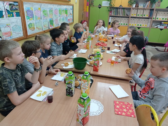 14 uczniów klasy II siedzi przy stołach pełnych ciasta, owoców i napojów.