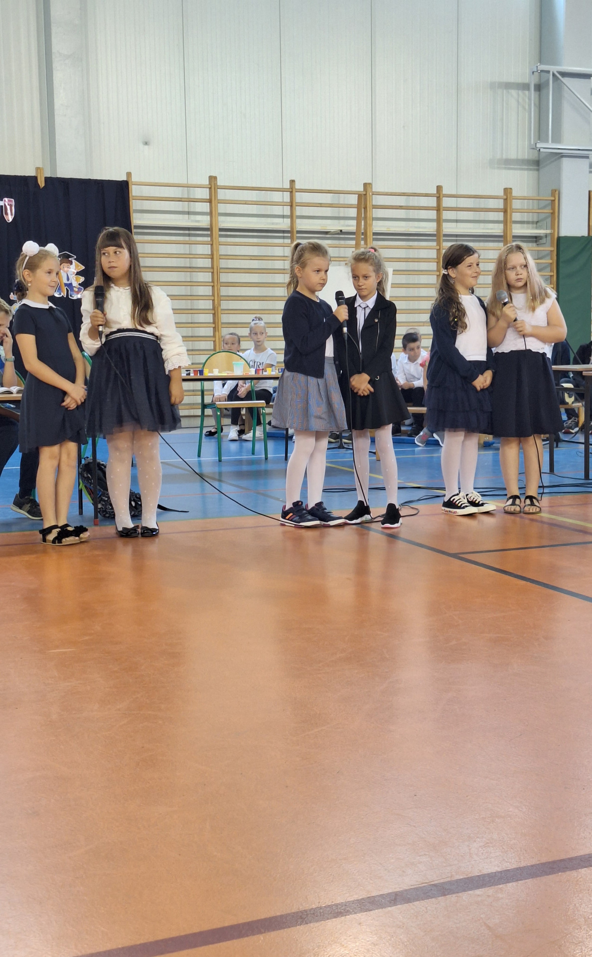 Grupa uczniów śpiewa piosenki na hali sportowej.