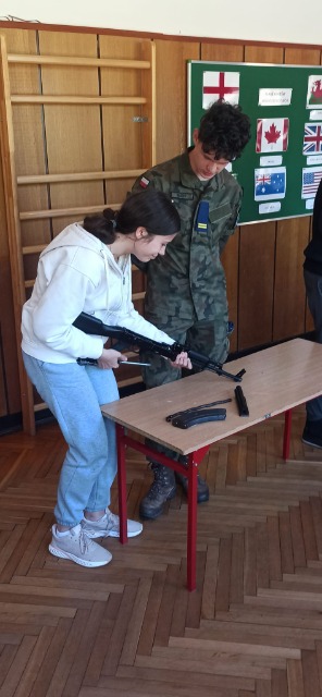Uczennica trzyma w ręku broń palną, obok niej stoi umundurowany chłopiec. Na stole, który stoi na przeciwko nich leżą elementy broni.