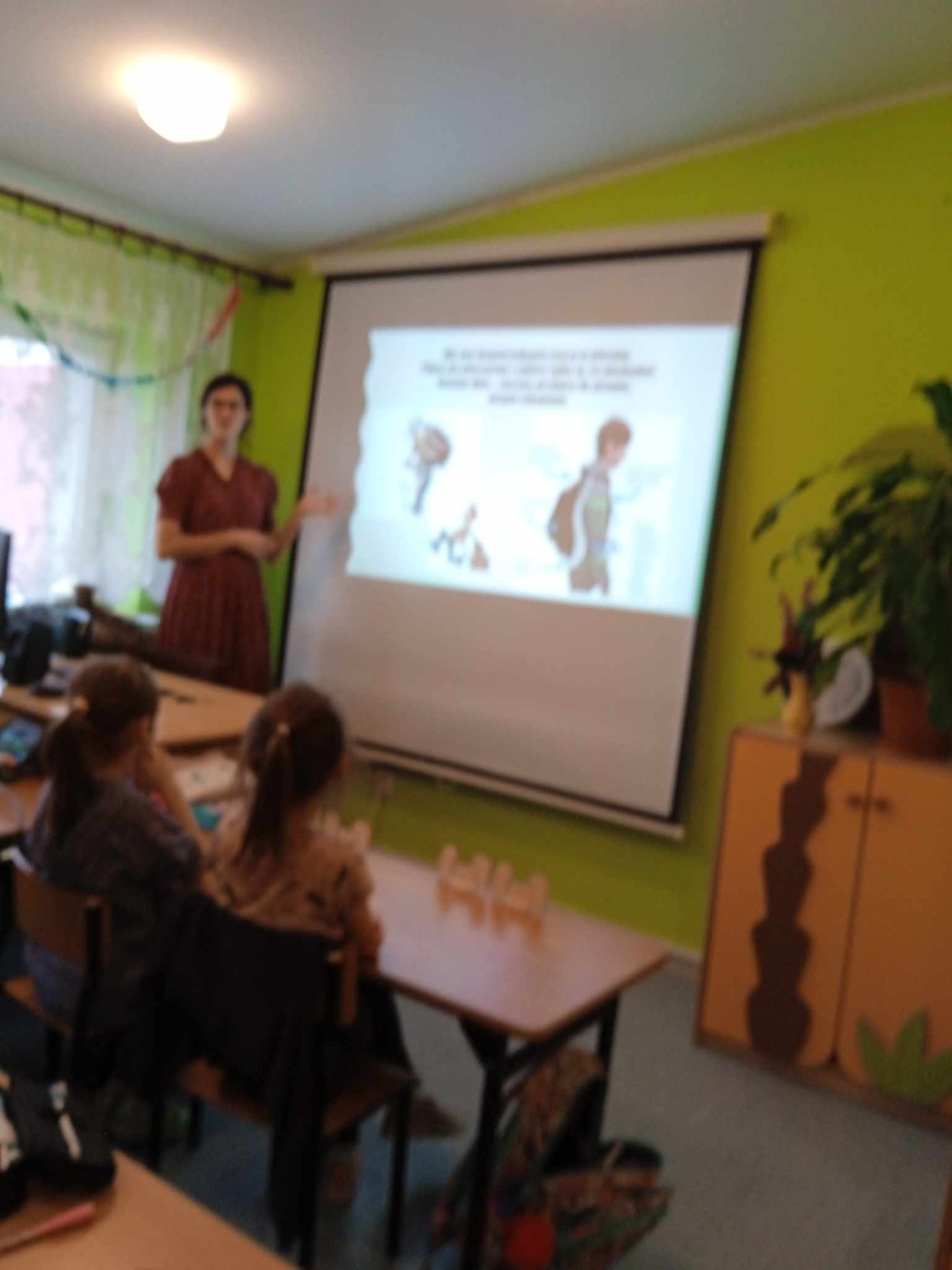 Nauczycielka stoi obok ekranu, na którym wyświetlane są slajdy z programu #ŻyjDobrze i opowiada uczniom o zasadach zdrowego zachowania.