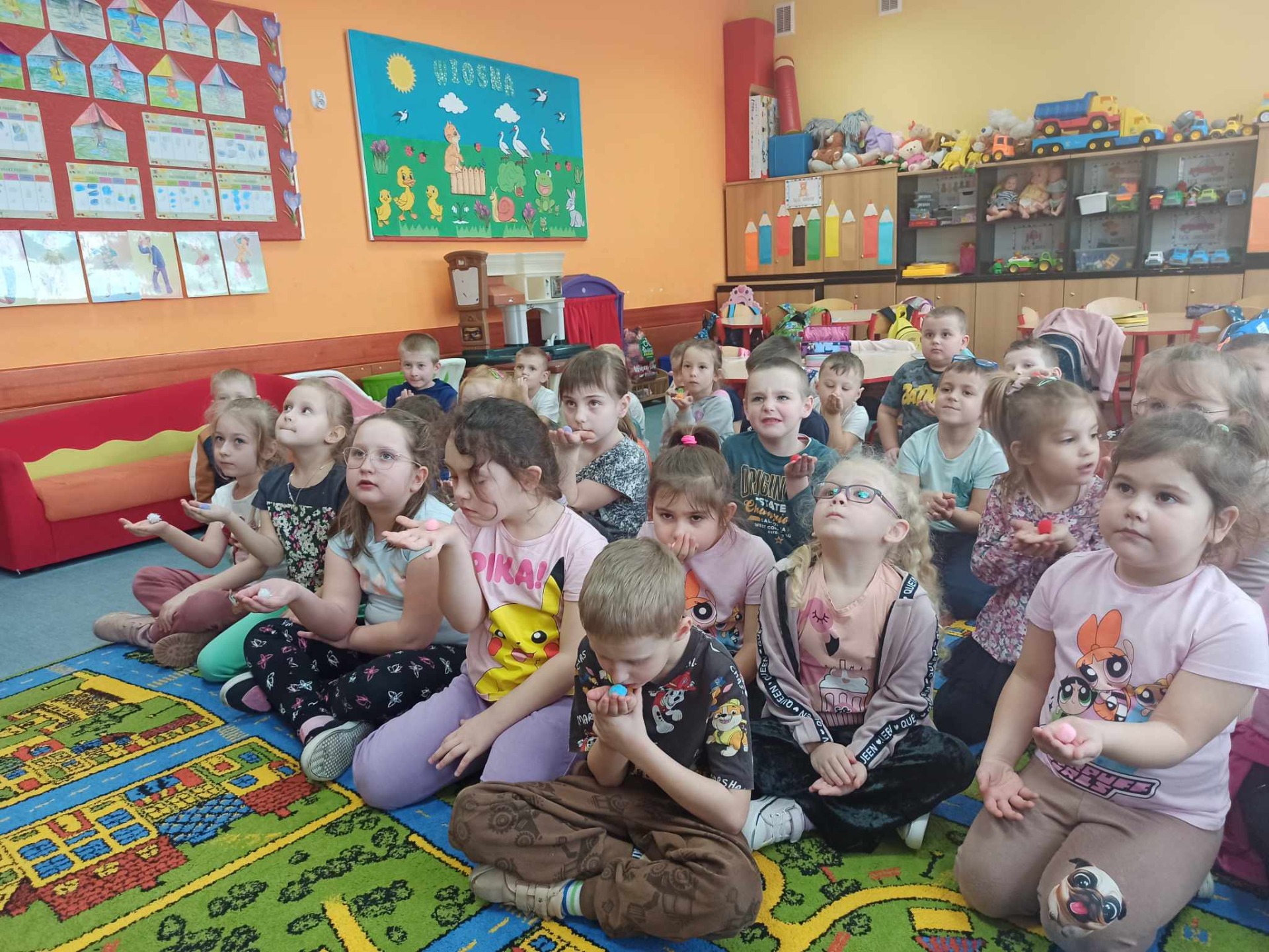 Kilkudziesięciu przedszkolaków siedzi na kolorowym dywanie i wykonuje ćwiczenia oddechowe.