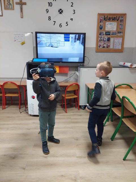 Dwaj chłopcy znajdują się w sali lekcyjnej. Jeden ma nałożone google, a drugi jest zwrócony twarzą do monitora.