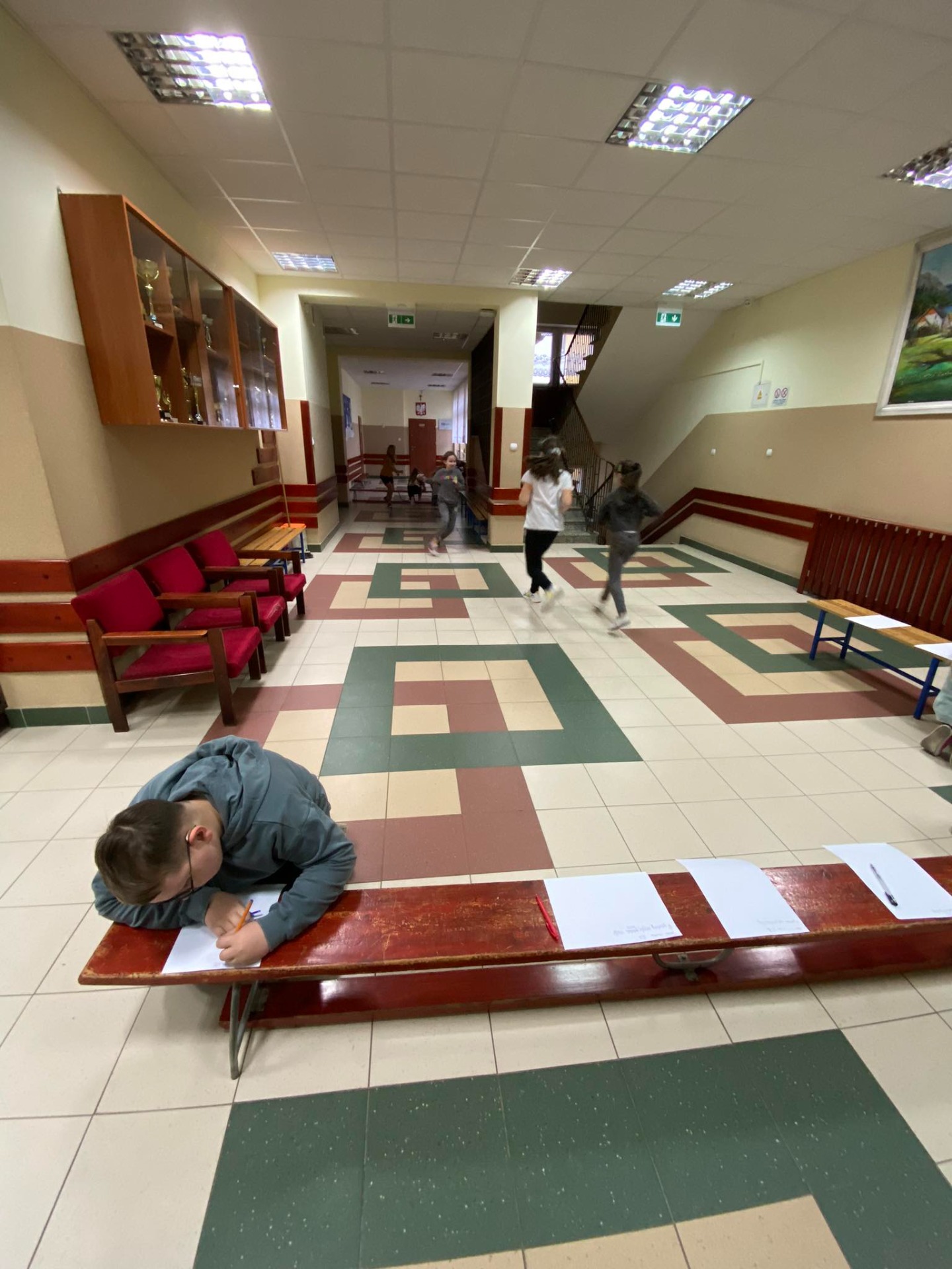 Uczniowie biegną po korytarzu do ławki, na której leżą kartki z tekstem