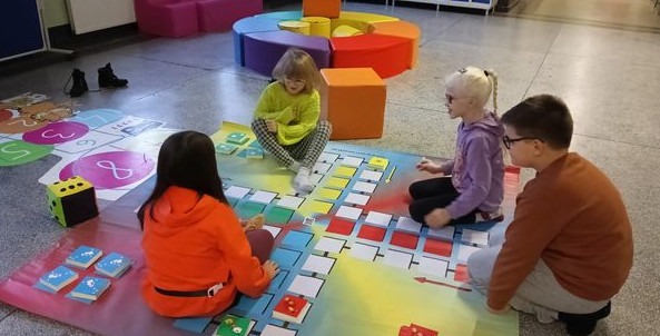 Kilkoro dzieci siedzi na podłodze i gra na rozłożonej kolorowej planszówce