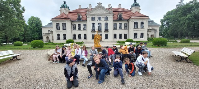 Grupa uczniów siedzi i kuca obok fontanny przy Pałacu Zamojskich w Kozłówce