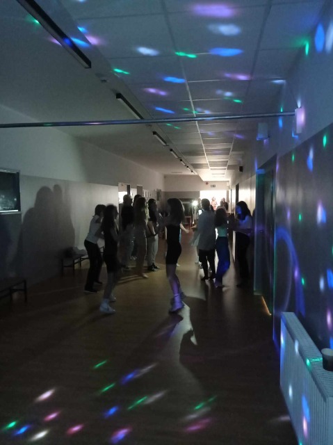 Na ciemnym tle korytarza szkolnego znajdują się tańczący uczniowie. W tle widać kolorowe światełka w odcieniach niebieskiego. 