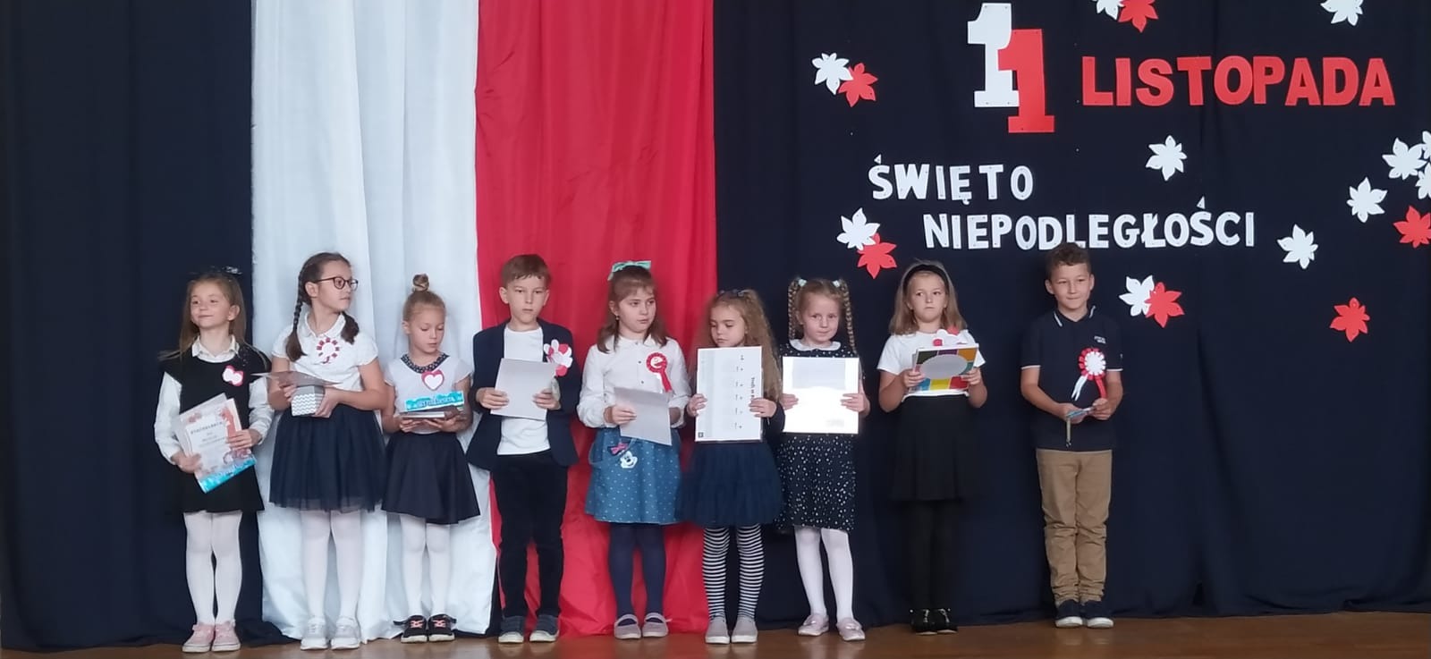 Nagrodzone dzieci stoją z dyplomami na tle dekoracji z okazji Dnia Niepodległości.