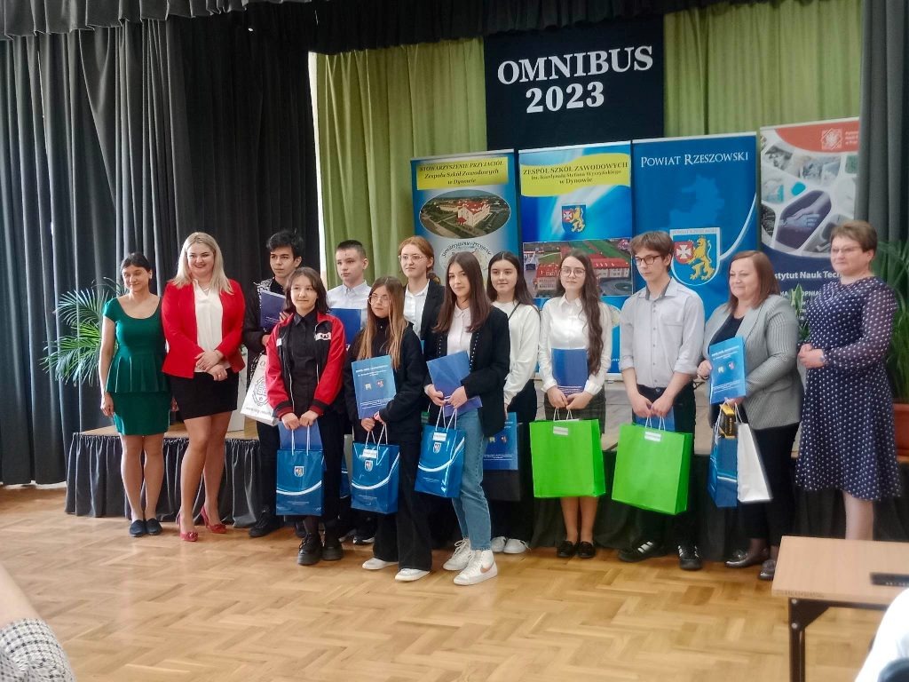 Na zdjęciu trzynastoosobowa grupa, w tym uczestnicy konkursu OMNIBUS 2023 -Język angielski (trzy dziewczyny z przodu i dziesięć osób z tyłu) oraz organizatorzy stoją przy scenie, w tyle na zielonym tle plakaty i napis "OMNIBUS 2023".