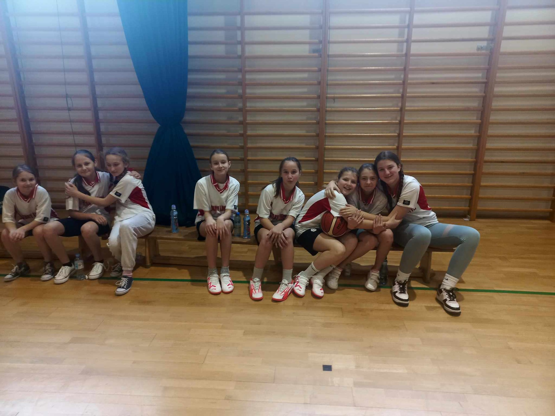 Grupa dziewcząt siedzi na ławce w  hali sportowej.